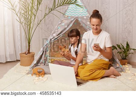 Image Of Woman And Her Daughter Wearing White T Shirts Sitting In Wigwam In Front Of Laptop, Kid Pla