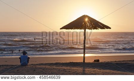 Beach Ocean Shoreline Man  Silhouetted Sitting Next To Wood Umbrella Watching Sunrise On The Sea Hor