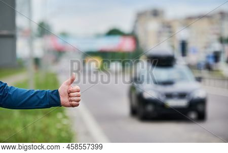 Close Up Of Mans Hand Hitchhiking By Roadside. Male Hand Showing Thumbs Up Gesture Outdoors On Blurr
