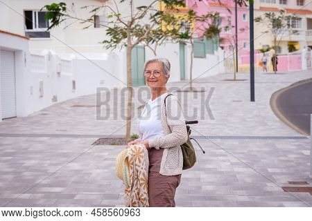 Beautiful Woman Traveler Walking In A City Street Holding Backpack. Smiling Elderly Caucasian Lady E