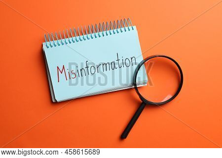 Notebook With Word Misinformation And Magnifying Glass On Orange Background, Flat Lay