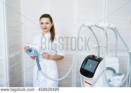 Lovely Woman Beautician Stands At Cosmetic Apparatus And Holds A Cellulite Nozzle In Her Hands. Youn