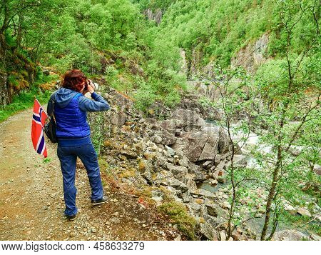 Tourist Woman With Norwegian Flag And Camera Walking Trough Mountains Path In Allmannajuvet Area Sau