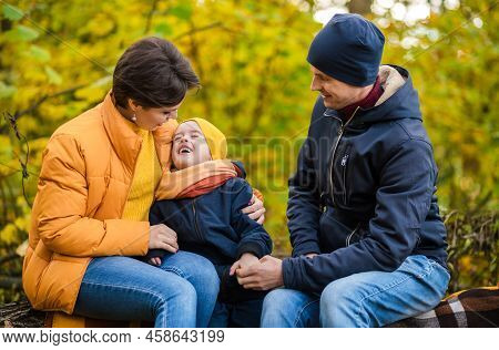 Mother, Father And Little Son Sit On A Plaid On A Log In The Autumn Park And Spend Time Together. Ho
