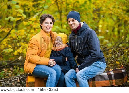 Mother, Father And Little Son Sit On A Plaid On A Log In The Autumn Park And Spend Time Together. Ho
