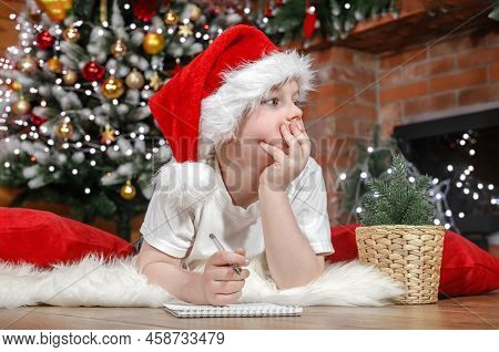 Happy Christmas Kids. Boy Child In Santa Claus Hat Writes A Letter To Santa Under The Christmas Tree