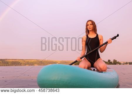 A Beautiful Girl Is Floating On A Supboard On A River. Young Woman Supsurfing At Sunrise.