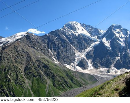 View Of Mount Cheget From The Village Of Terskol. A High Mountain With Snowy Peaks Against A Blue Sk