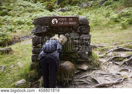 Ermida (portugal), June 26, 2022. Stone Fountain. Woman Drinking Water In A Forest, Near The Waterfa
