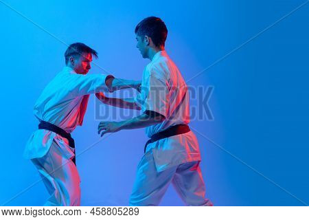 Studio Shot Of Sports Training Of Two Karatedo Fighters In Doboks Isolated On Blue Background In Neo