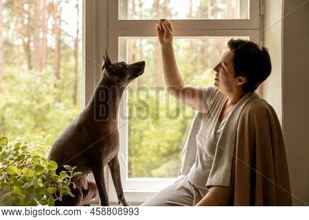 Middle Age Beautiful Woman Sitting On Windowsill With Her Dog. 50-year-old Woman Spending Time With 