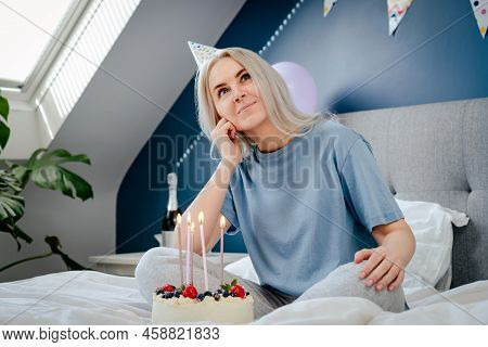Thoughtful Smiling Woman In Birthday Party Cap With Festive Cake With Candles On The Bed In Decorate