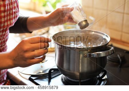 Woman Adding Salt To Boiling Water In Pot On Stove Indoors, Closeup