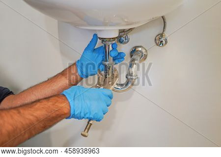 Plumber With Blue Gloves Repairing Sink Siphon In Kitchen. Plumber Repairs And Maintains Chrome Siph