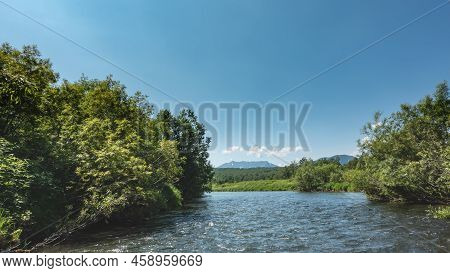 The Riverbed Bends Between The Green Banks. Lush Vegetation Near The Water. Mountains Against The Bl