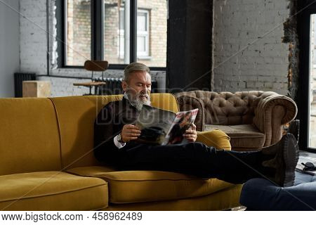 Elderly Focused Caucasian Man Reading Journal On Sofa At Home. Trendy Bearded Pensioner Wearing Casu