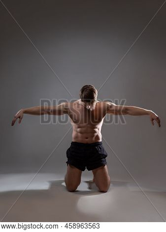 Muscular Man In An Artistic Pose, Portrait On A Gray Background. The Athlete Guy Poses On His Knees 