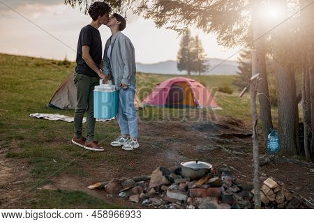 Male Tourist Kissing Girlfriend And Holding Portative Fridge At Camping Site. Rural Rest.