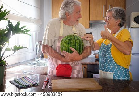 Cheerful Senior Caucasian Couple In Home Kitchen Holding A Big And Heavy Watermelon  - Hidration, Fr