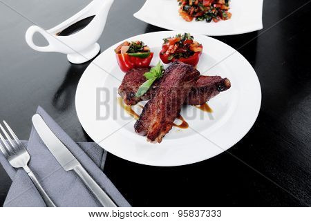 fresh red beef meat steak barbecue garnished vegetable salad and basil on white plate over black wooden table with bbq sauce in sauceboat