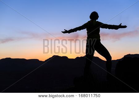Woman hiking silhouette in mountains sunset happiness. Female hiker climber or trail runner with arms outstretched on mountain top looking at beautiful night sunset inspirational landscape.