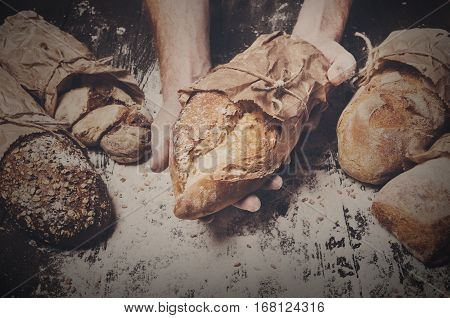 Warm fresh rye whole grain bread, wrapped in craft paper. Healthy baking and cooking concept background. Hands hold loaf on wooden table background, sprinkled with flour. Soft toning, filtered