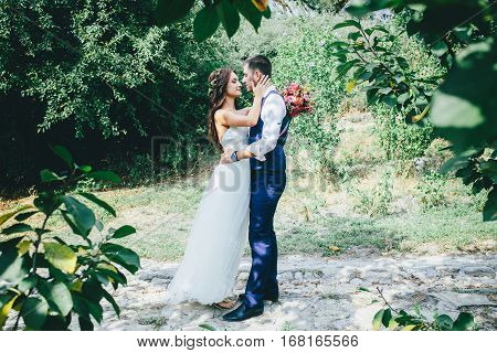 happy groom in a dark blue suit hugs a beautiful bride in a white wedding dress with pink and purple bouquet of flowers outdoors