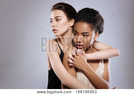 Looking in the same direction. Glamorous well groomed tender women wearing delicate dresses and working during a photoshoot in a studio while one of them hugging anther