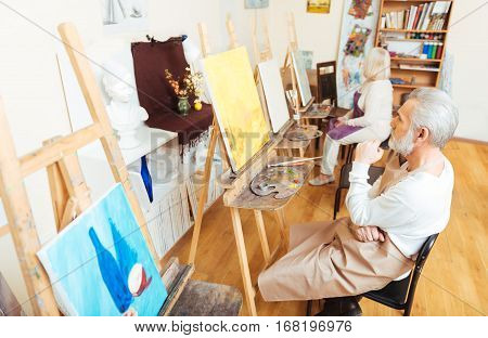 For inspiration. Elderly inspired thoughtful artist sitting in painting class and looking at painting while enjoying his hobby