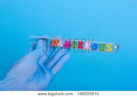 Word dangerous of colored wooden letters in test-tube. Hand in glove holding a test tube on a blue background