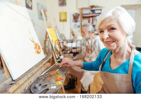 Having good time. Female elderly delighted artist smiling and painting picture while working in painting studio.