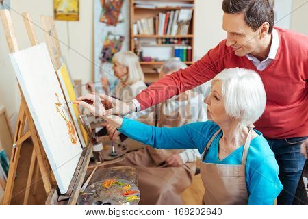 Give an advice. Delighted professional artist helping elderly woman while spending time in painting studio and drawing a picture.