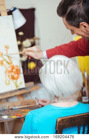Be good. Professional young concentrated artist teaching elderly woman while drawing and spending time in painting school.