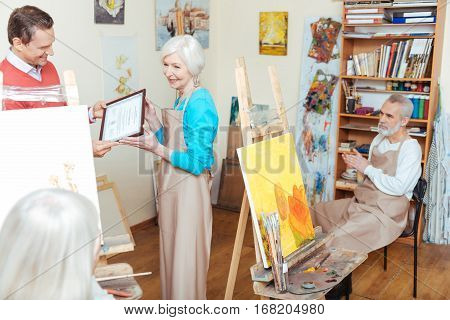 Take it. Handsome young delighted man giving artists diploma to woman while spending time in painting school and working as a teacher.