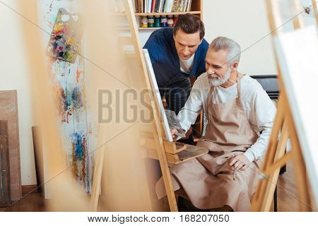 Good lesson. Glad handsome young artist helping elderly man while he painting and spending time in painting studio.