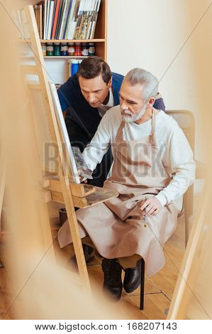 Pleasant lesson. Talented handsome young artist helping elderly man while he painting and spending time in painting studio.