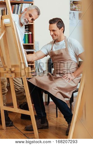 Hardworking colleagues. Concentrated elderly professional man painting while working with his young colleague and spending time in painting studio.