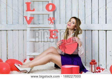 Blonde girl with blue eyes portrait. Opening a gift box. Romantic scene. Red hearts and lights. Red and White background