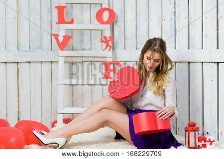 Blonde girl with blue eyes portrait. Opening a gift box. Romantic scene. Red hearts and lights. Red and White background