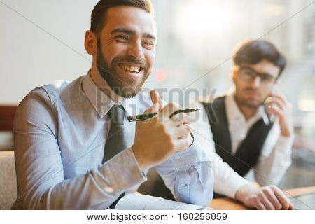 Portrait of young attractive company executive sitting at table with clasped hands, holding pen and smiling at camera cheerfully, sitting next to his pensive assistant.