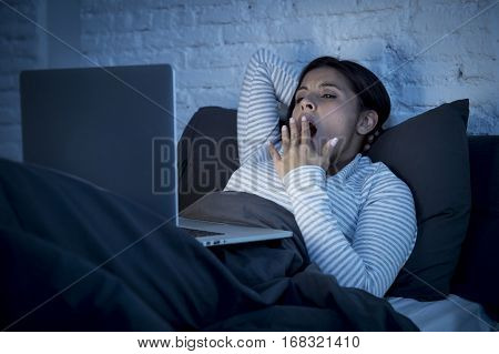 young beautiful hispanic internet addict woman in pajamas on bed at home bedroom working bored and tired with laptop computer late at night in dark room light yawning exhausted falling sleep