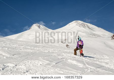 Stylishly dressed snowboarder stands and walks on mountain slopes of an extinct volcano Elbrus on a background of pure blue sky