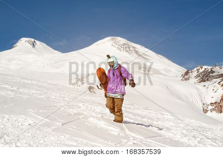 Stylishly dressed snowboarder stands and walks on mountain slopes of an extinct volcano Elbrus on a background of pure blue sky