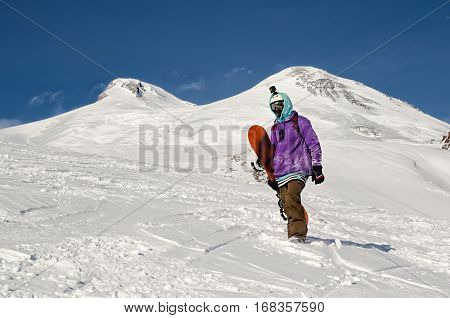 Stylishly dressed snowboarder stands and walks on mountain slopes of an extinct volcano Elbrus on a background of pure blue sky