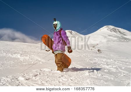 Stylishly dressed snowboarder stands and walks on mountain slopes of an extinct volcano Elbrus on a background of pure blue sky