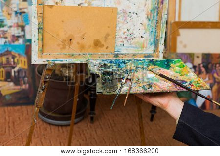 Artist hand holding palette with mixed pigment and paintbrushes ready to paint on canvas.