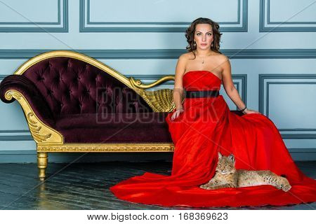 Woman in red dress sits on couch, lynx cub lies on dress hem on floor.