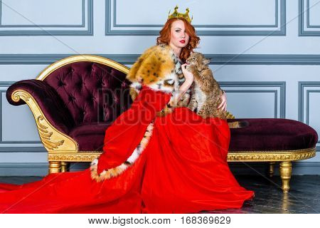 Red-haired woman in red dress, cloak and with crown on head sits on couch holding lynx cub.