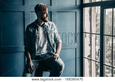 Young and confident. Handsome young man in light blue jeans shirt looking in window while sitting on chair