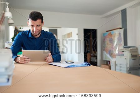 Confident and successful businessman wearing an open collar shirt sitting at his desk using a digital tablet in his modern office